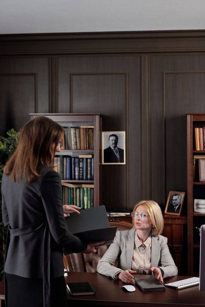 Two businesswomen at a meeting in a book-filled office. Professional and focused setting.